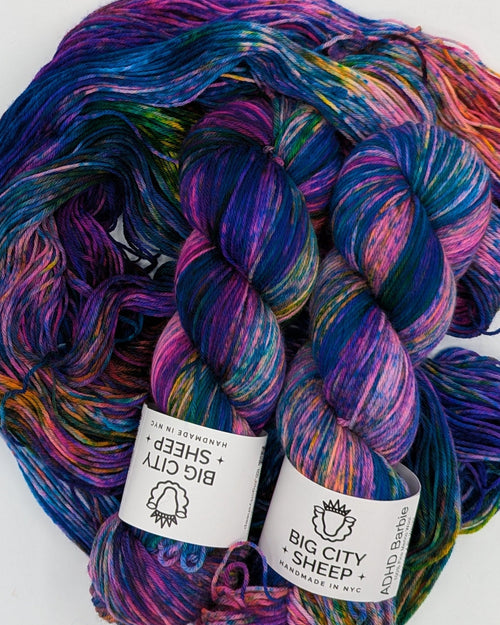 A skein of variegated yarn with purple, blue, pink, and orange tones, displayed on a white sheet with two white paper labels attached.
