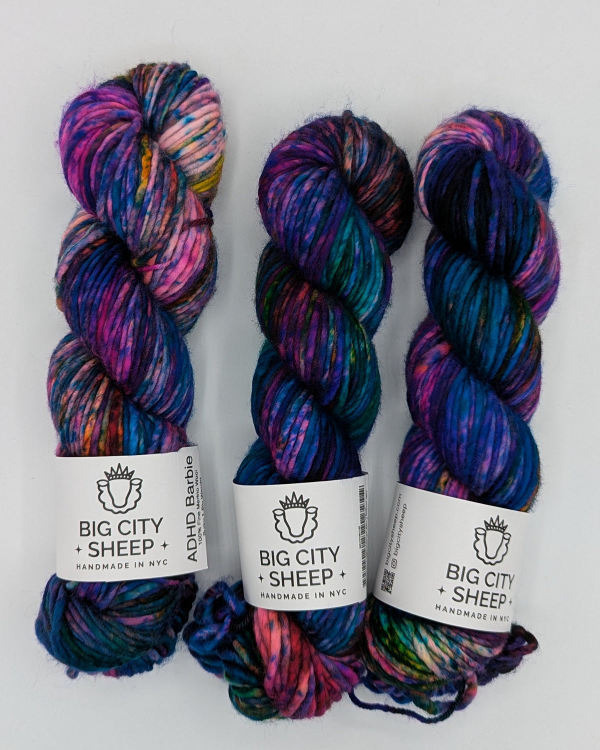 Three skeins of variegated yarn in purple, blue, pink, and green hues, each with a white paper label reading 'BIG CITY SHEEP', arranged on a white sheet of paper.
