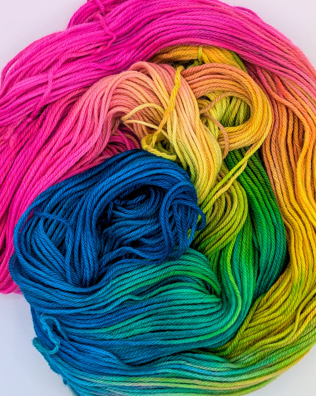 A close-up of a skein of variegated yarn showing a smooth rainbow gradient transition from blue and green to yellow, orange, and bright pink, photographed on a clean white background.