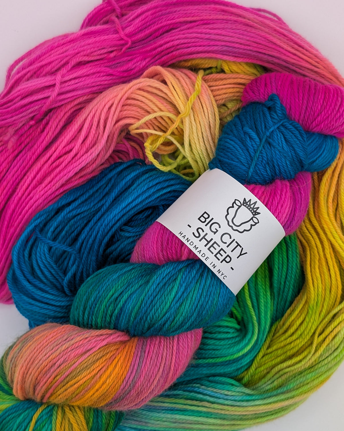 A close-up of a skein of variegated yarn with multiple bright colors including pink, blue, green, yellow, and orange, wrapped with a white paper label that reads 'BIG CITY SHEEP- HANDMADE IN NYC.' on a plain white background.