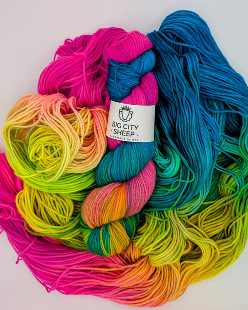 A pile of colorful yarn skeins in a rainbow gradient, with a white paper label reading 'BIG CITY SHEEP'