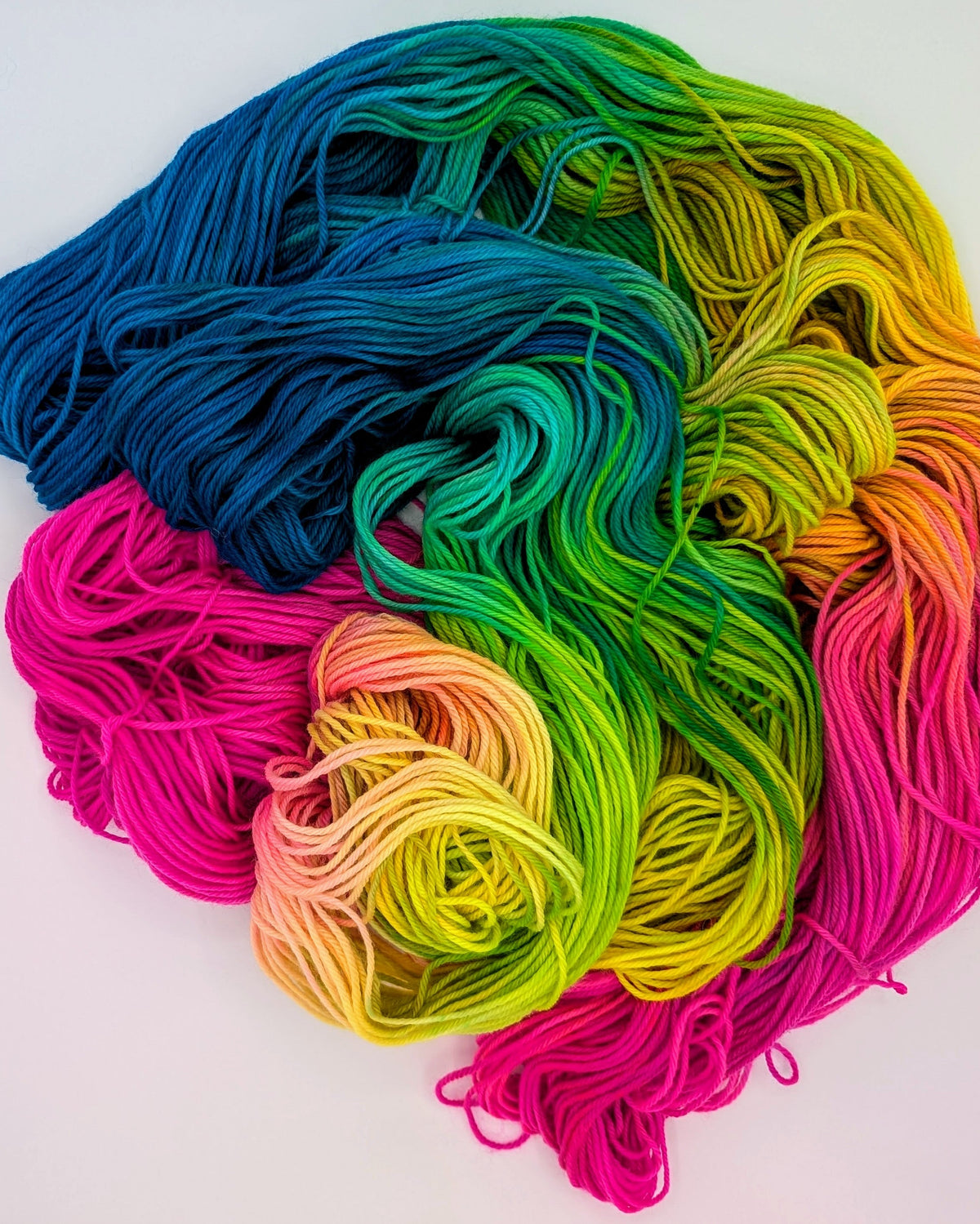 A pile of vibrant, multi-colored yarn skeins arranged in a rainbow gradient of blue, green, yellow, and pink on a plain white background.