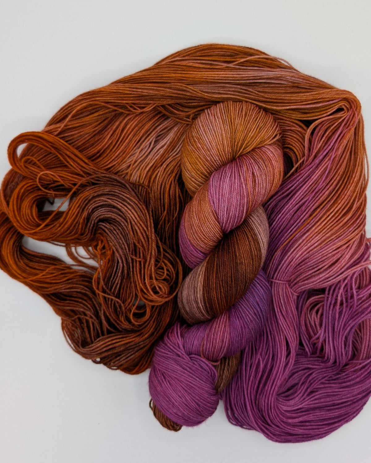 Two skeins of gradient yarn arranged on a plain white background. One skein is twisted and sitting on top of the other, which is open. The colors are rust orange, purple, and blend of brown and pink, showing a color-shifting or multi-tonal effect.