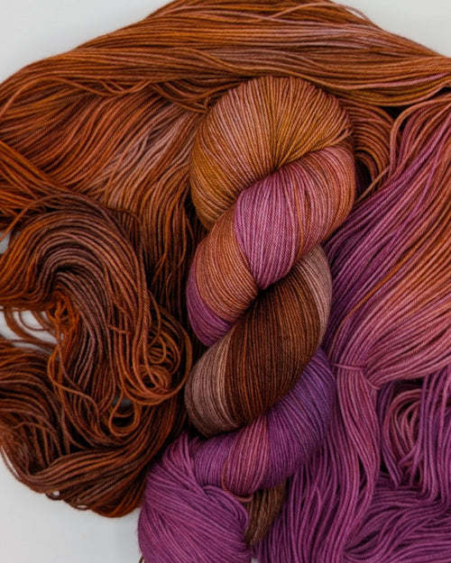 Two skeins of gradient yarn arranged on a plain white background. One skein is twisted and sitting on top of the other, which is open. The colors are rust orange, purple, and blend of brown and pink, showing a color-shifting or multi-tonal effect.