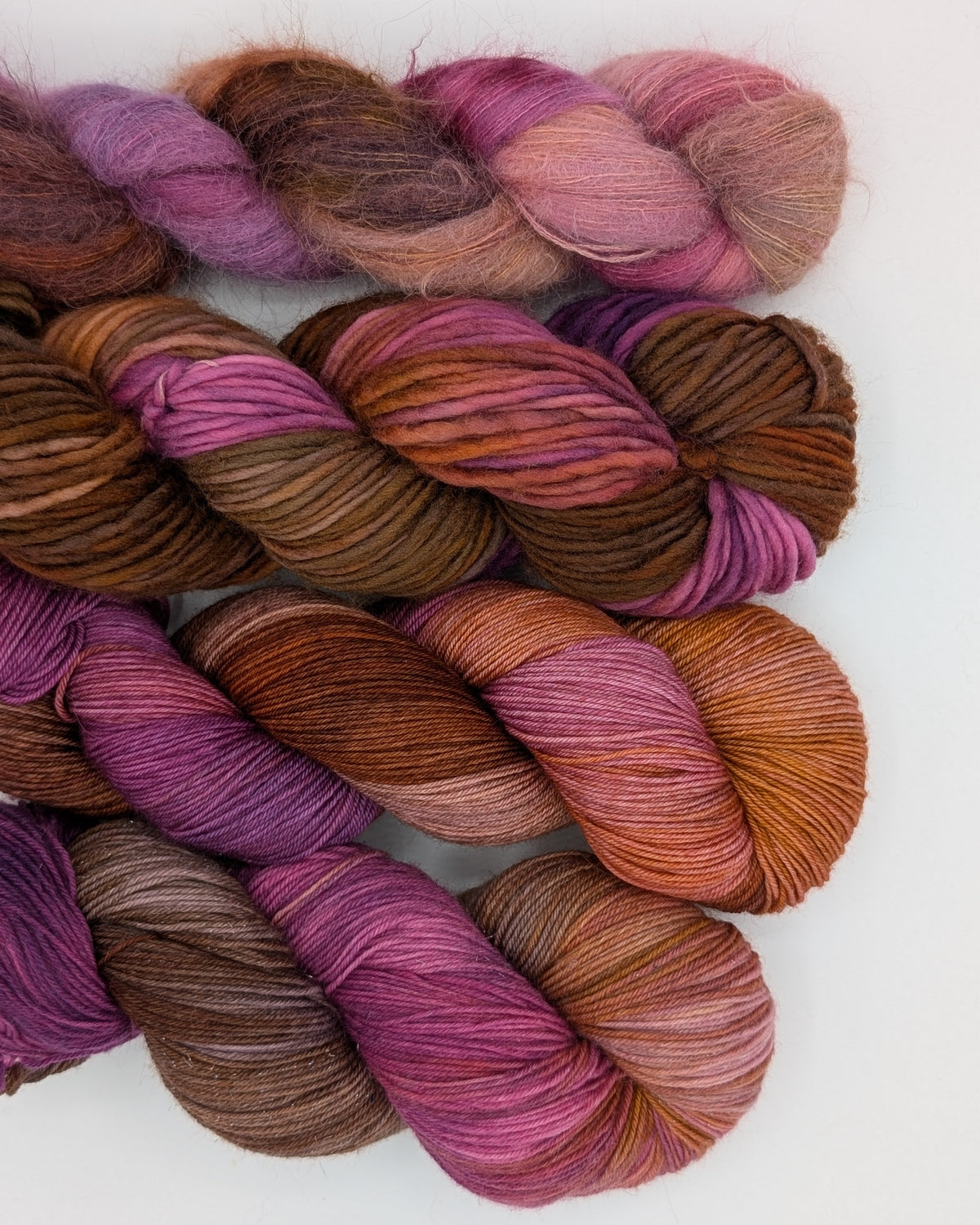 Four skeins of variegated yarn arranged side by side on a plain white background. The yarn displays a color-shifting variegated pattern blending shades of purple, pink, brown, and orange. In order from left to right, they are sock, fingering, bulky and alpaca. 