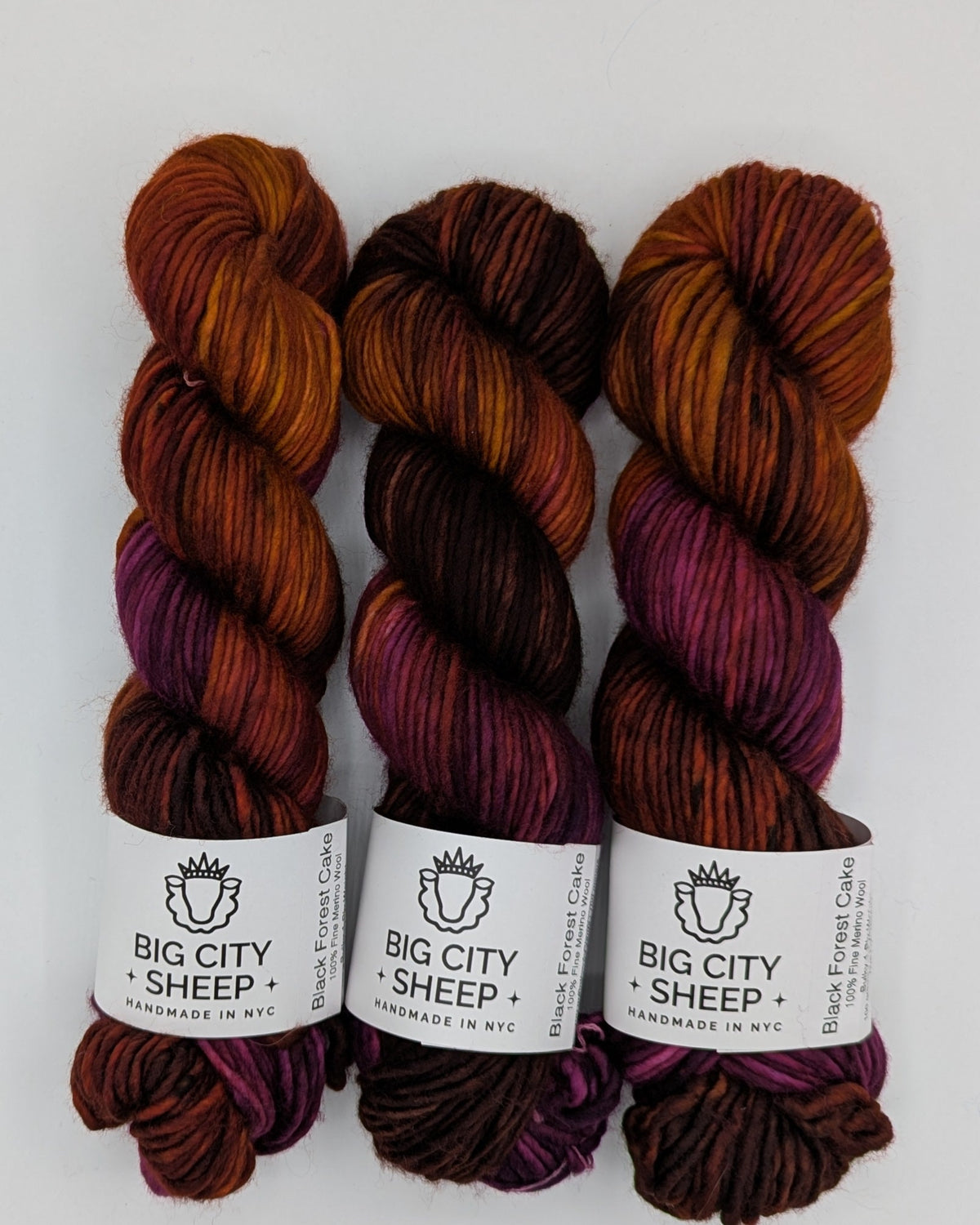 Three skeins of yarn from Big City Sheep, featuring a marbled colorway of rust orange, deep burgundy, and plum purple, arranged on a white surface.
