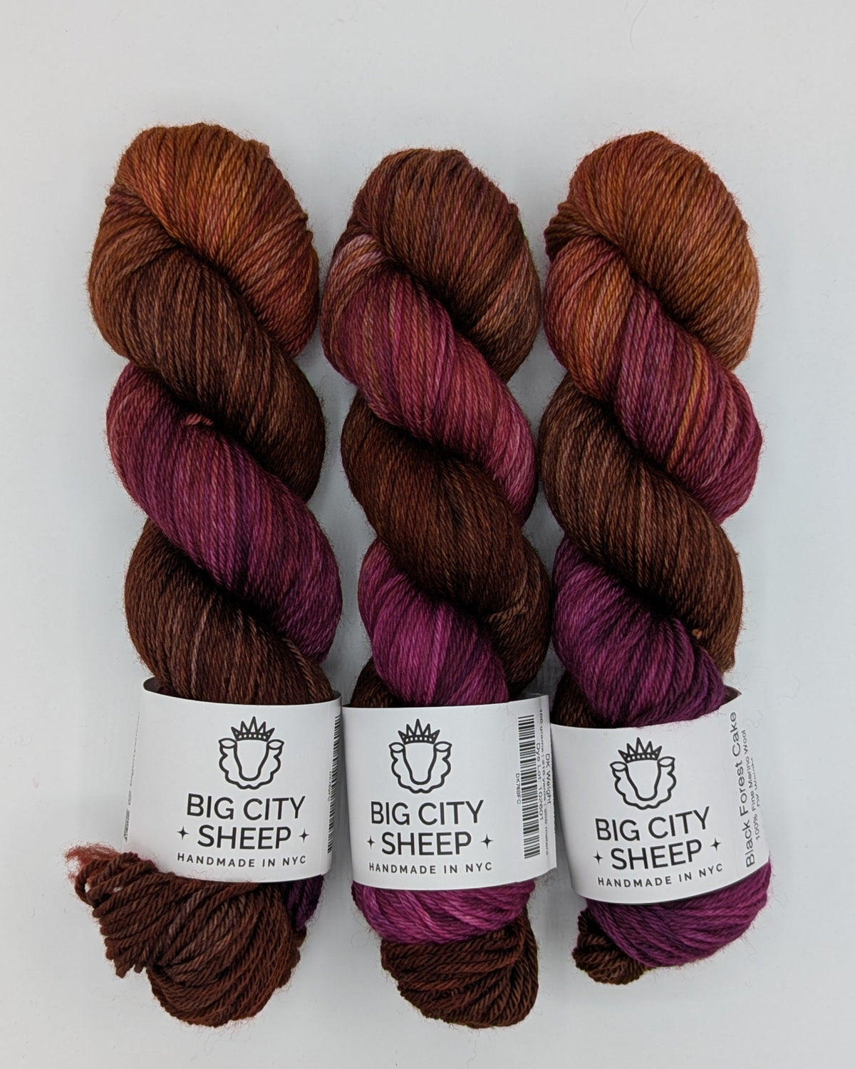Three skeins of yarn from Big City Sheep, featuring a variegated colorway of rust brown, deep burgundy, and plum purple, each with a white paper label, displayed on a white background.