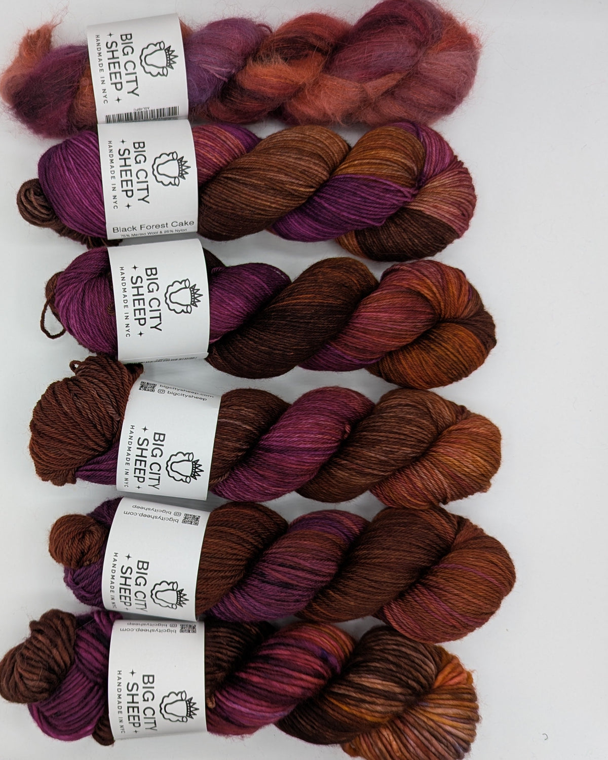 Six skeins of variegated yarn arranged vertically on a white surface. The yarn transitions through shades of rust orange, deep burgundy, plum purple, and dark brown.
