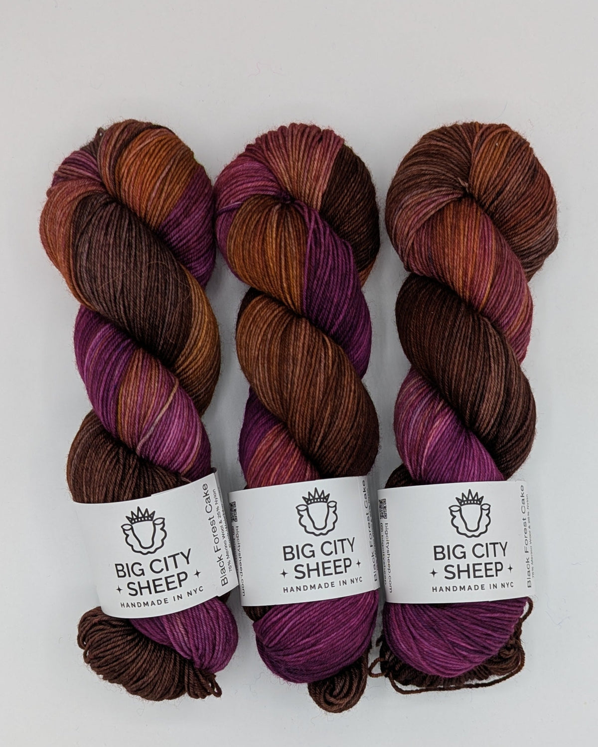 Three skeins of variegated yarn in shades of purple, brown, and orange, each with a white paper label reading 'BIG CITY SHEEP', arranged on a white surface.