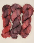 Three twisted hanks of variegated yarn in shades of burgundy, red, and black, arranged side-by-side on a light beige background.