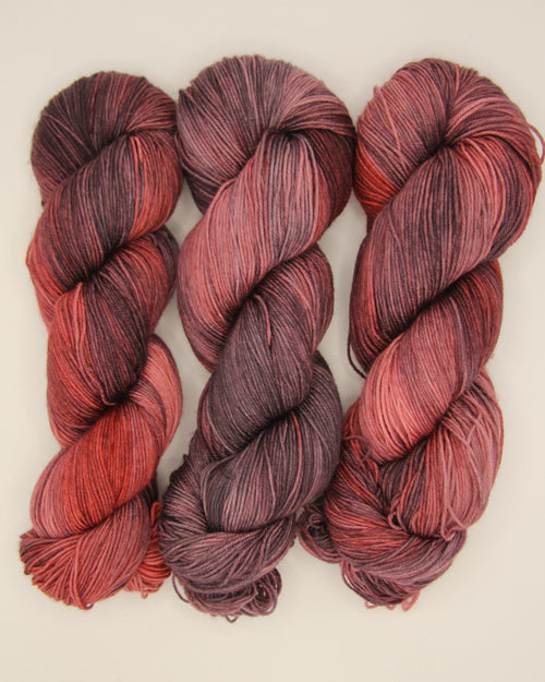 Three twisted hanks of variegated yarn in shades of burgundy, red, and black, arranged side-by-side on a light beige background.