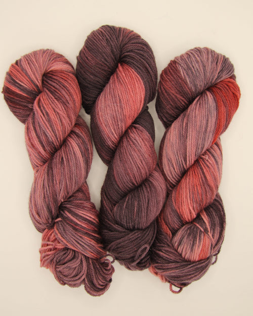 Three skeins of variegated yarn with a color-shifting pattern transitioning from deep burgundy and black to warm rust red, displayed on a clean off-white background.