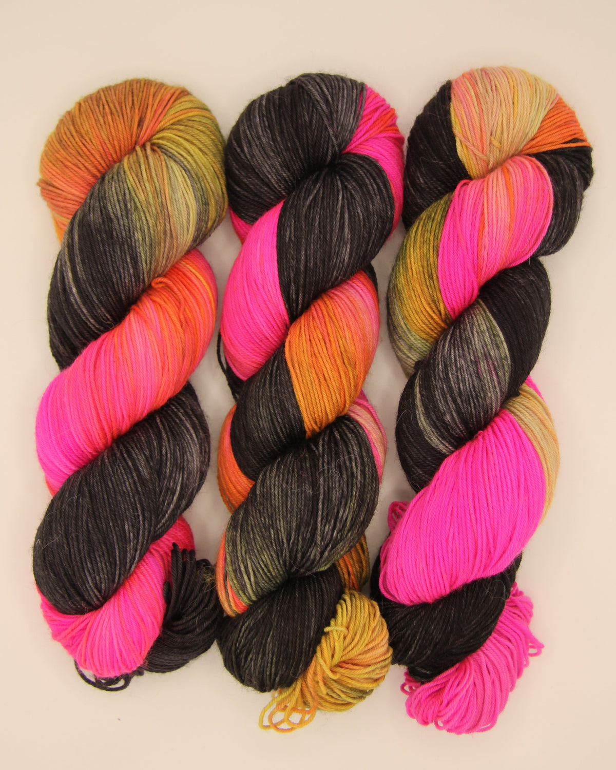 Three twisted hanks of variegated yarn displayed side-by-side on a plain, light beige background. The yarn features twisted sections of black, orange, hot pink, and yellow.