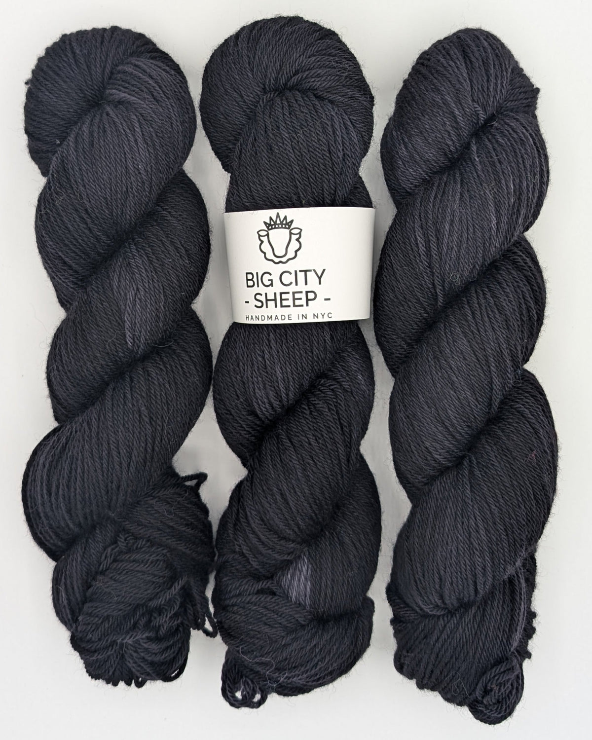 Three skeins of dark charcoal gray yarn with a white paper label in the center that reads 'BIG CITY SHEEP - HANDMADE IN N.Y.