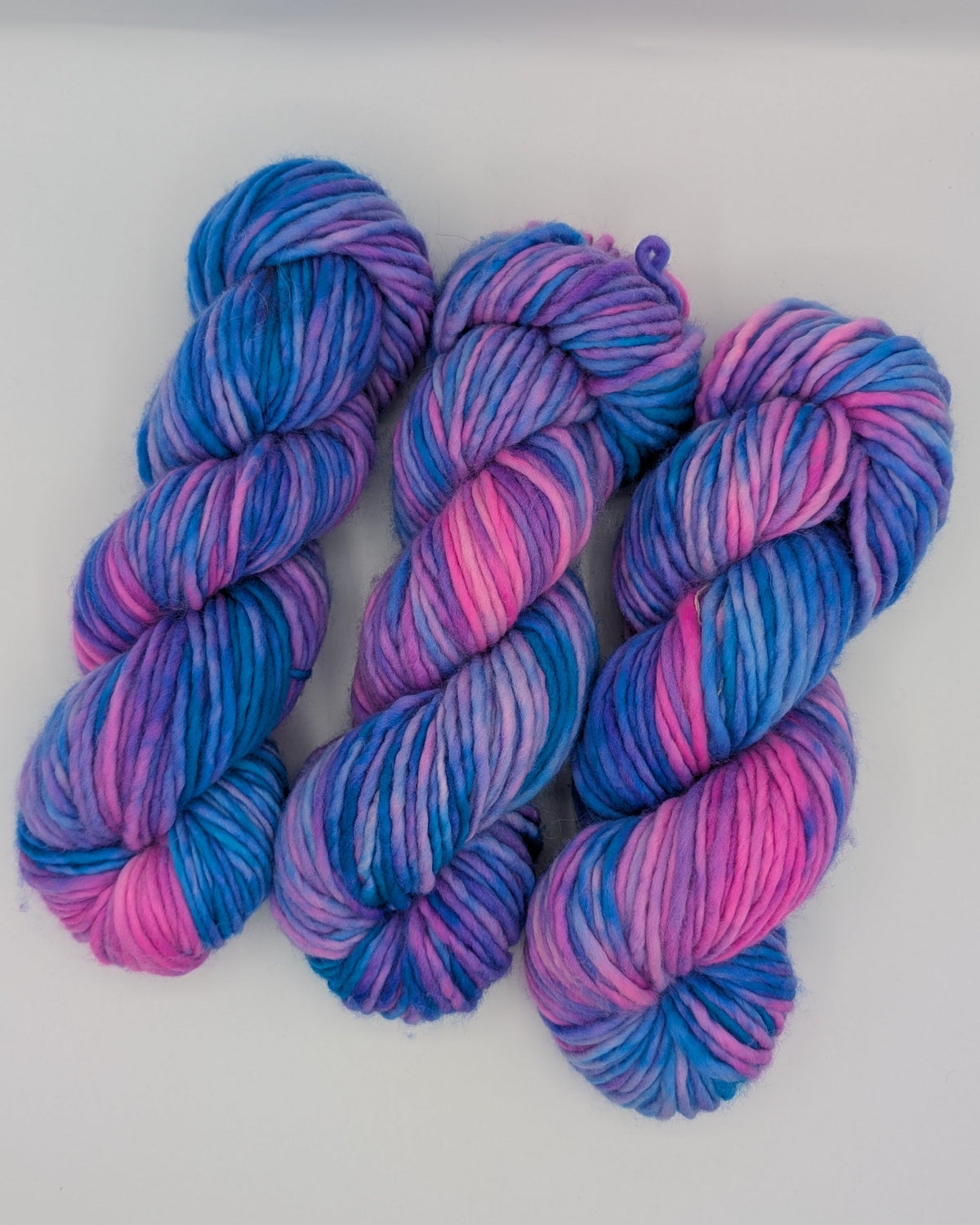 Three hanks of chunky variegated yarn in shades of blue, purple, and pink, arranged side-by-side on a plain white surface.