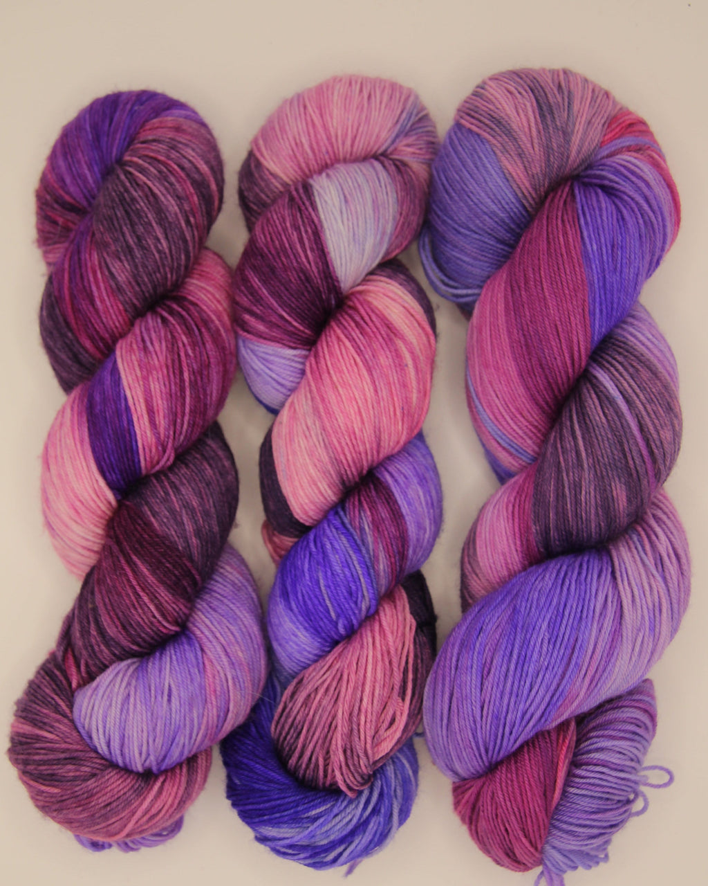 Three hanks of variegated yarn in shades of purple, pink, and magenta, arranged side by side on a light beige background.