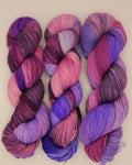 Three hanks of variegated yarn in shades of purple, pink, and magenta, arranged side by side on a light beige background.