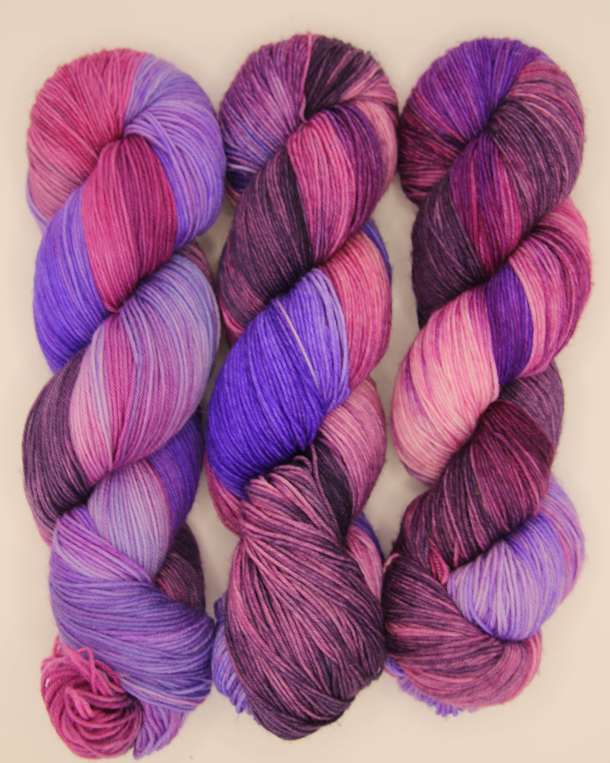 Three hanks of variegated yarn in shades of purple, pink, and magenta, arranged side-by-side on a light beige background.