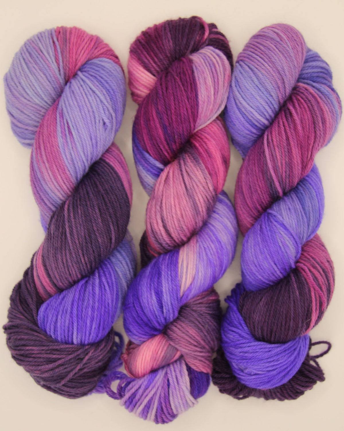 Three skeins of variegated yarn in shades of purple, pink, and violet, arranged horizontally on a light beige background.
