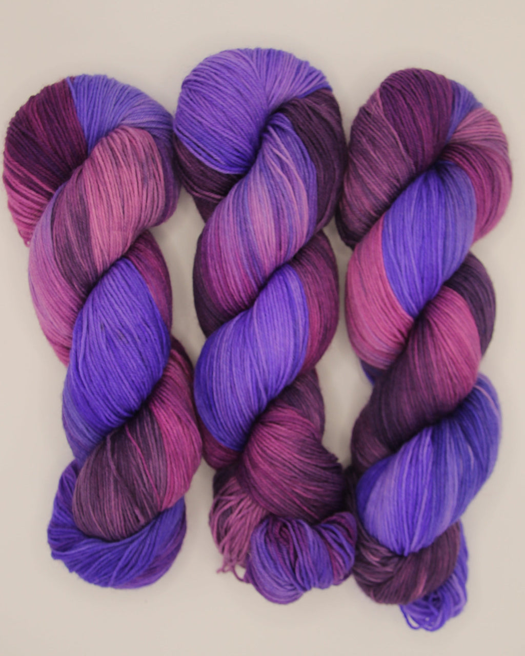 Three hanks of variegated yarn in shades of purple, violet, and magenta pink, arranged side-by-side on a plain off-white background.