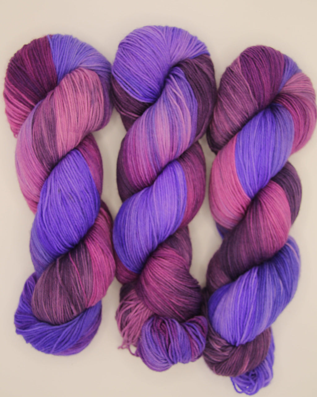 Three hanks of variegated yarn in shades of purple, violet, and magenta pink, arranged side-by-side on a light beige background.