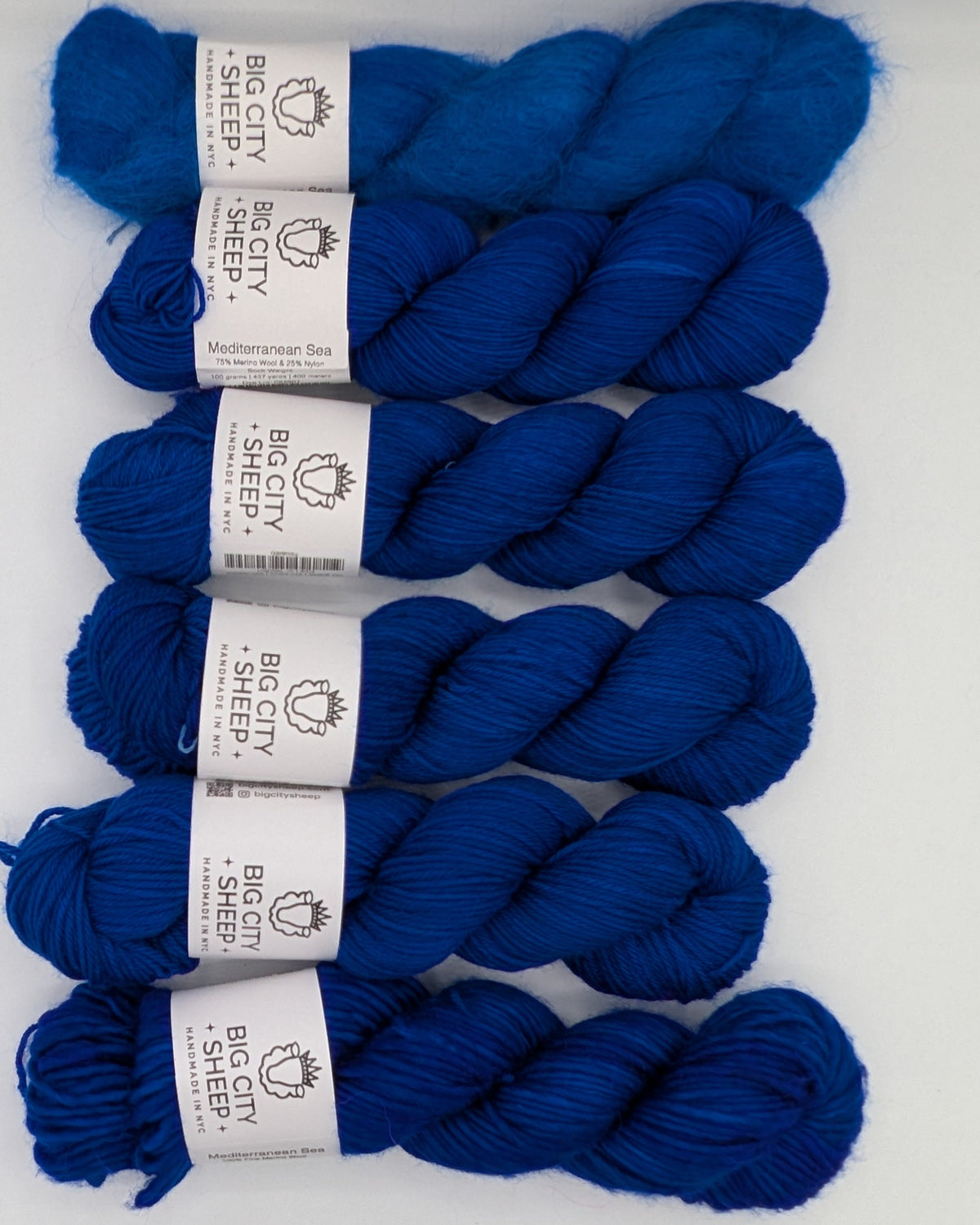 Six skeins of deep blue yarn arranged vertically in a clear plastic box lined with white paper. Each skein has a white paper label wrapped around it.