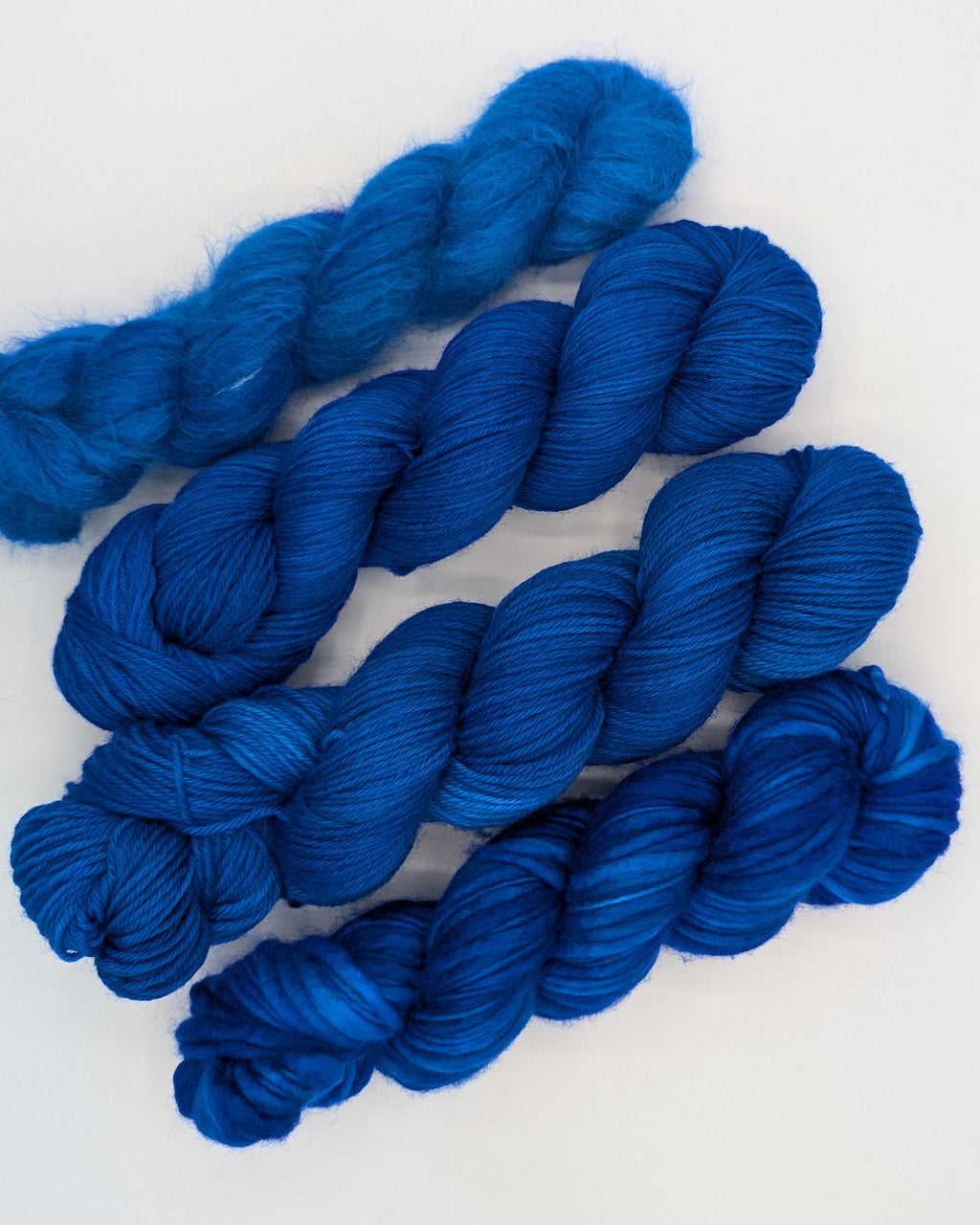 Four skeins of vibrant royal blue yarn arranged diagonally on a clean white background. In different weights, from left right: alpaca silk lace, fingering, worsted, chunky slub.