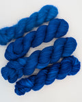 Four skeins of vibrant royal blue yarn arranged diagonally on a clean white background. In different weights, from left right: alpaca silk lace, fingering, worsted, chunky slub.