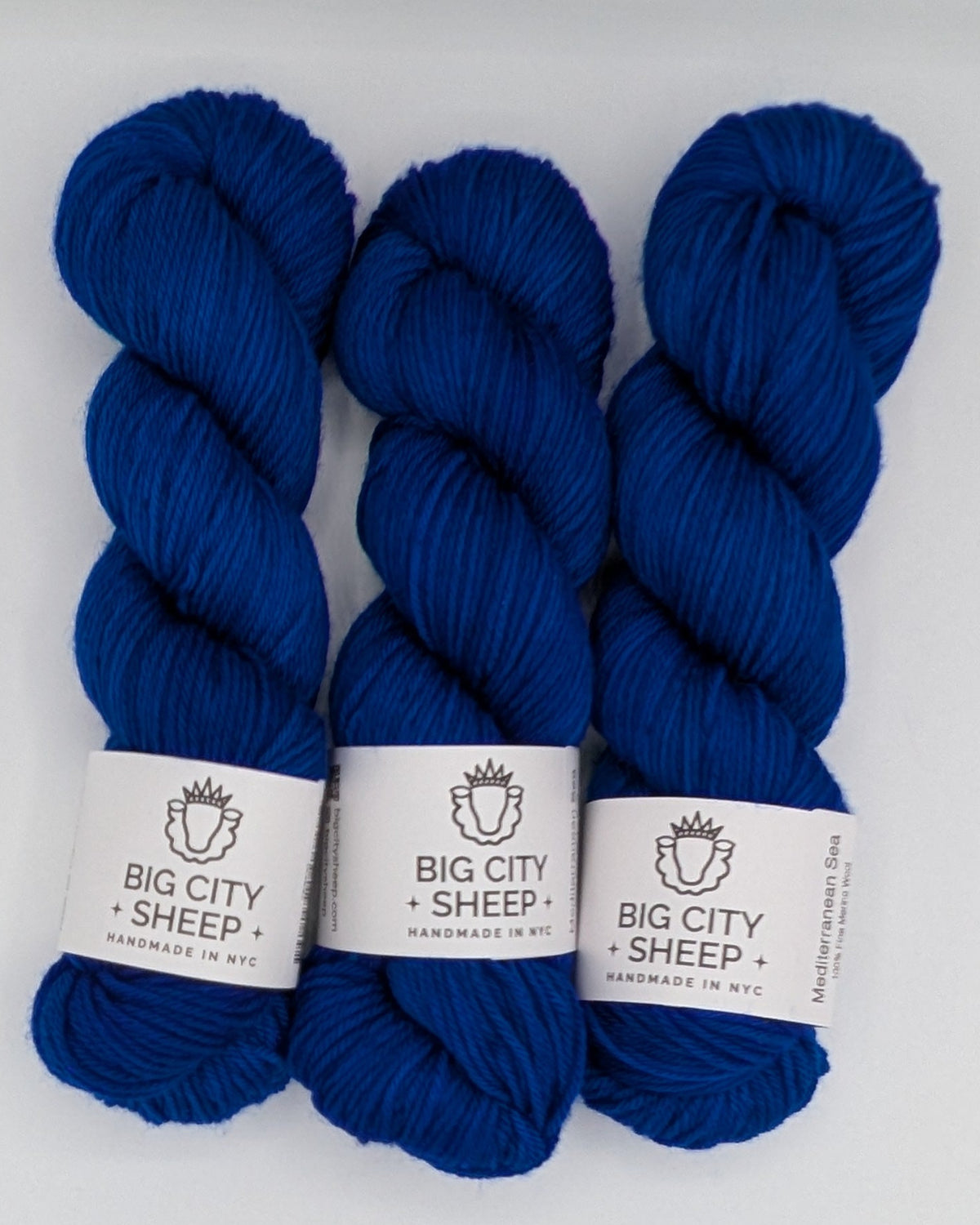 Three skeins of royal blue yarn with white paper labels reading 'BIG CITY SHEEP' arranged on a white surface inside a textured box.