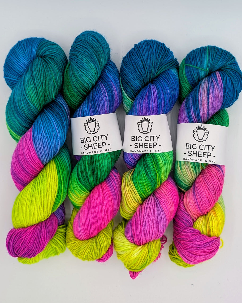 Four hanks of variegated yarn arranged side by side on a white background. Each hank features a blend of bright blue, teal, green, pink, and yellow, with a white paper label in the center reading 'BIG CITY SHEEP - Handmade in NY'.