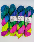 Four hanks of variegated yarn arranged side by side on a white background. Each hank features a blend of bright blue, teal, green, pink, and yellow, with a white paper label in the center reading 'BIG CITY SHEEP - Handmade in NY'.