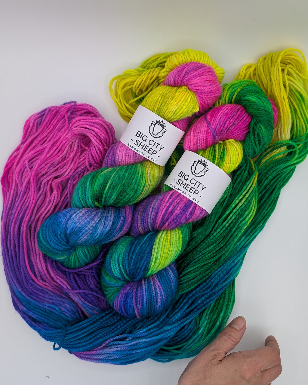 Two twisted skeins sit atop a third open skein of vibrant, multicolor variegated yarn from Big City Sheep, featuring bright shades of pink, purple, blue, green, and yellow, with a hand visible at the bottom right for scale.