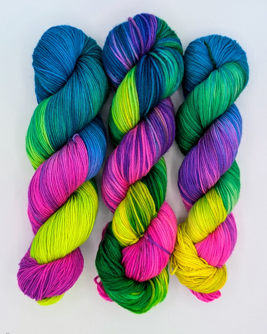 Three skeins of variegated yarn arranged side by side on a plain white background. The yarn features vibrant, blended colors including blue, teal, green, purple, pink, and neon yellow. From left to right sock, worsted, fingering. 