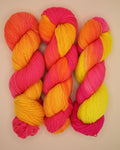 Four skeins of variegated yarn arranged in a grid on a light beige background. The yarn features a gradient color story transitioning from hot pink, to bright orange, and ending in vibrant yellow.