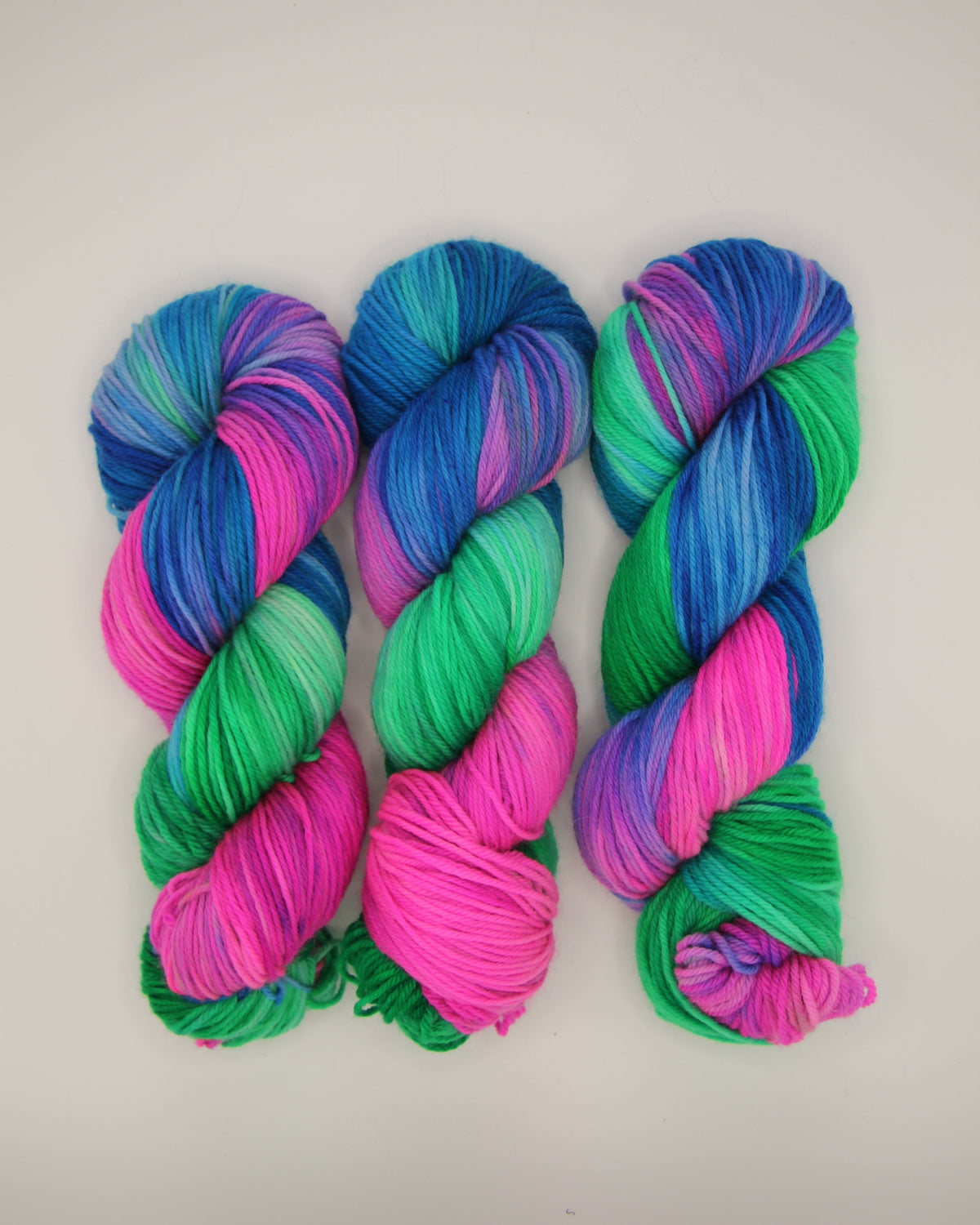 Three skeins of variegated yarn arranged vertically on a plain light background. The yarn displays a vibrant blend of bright pink, teal green, and blue with purple undertones.