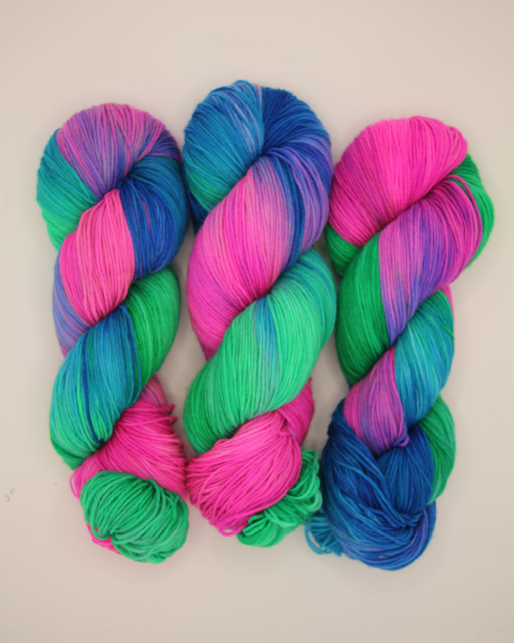 Three skeins of vibrant, variegated yarn arranged vertically on a plain off-white background. The yarn features a striking blend of bright pink, green, and blue in a twisted, variegated pattern.