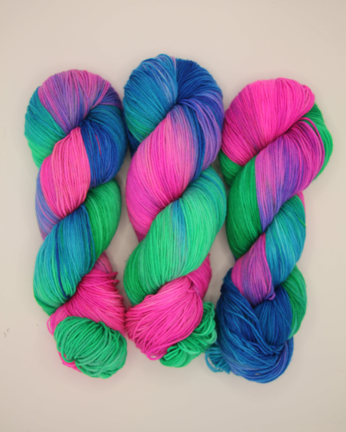 Three skeins of vibrant, variegated yarn arranged vertically on a plain off-white background. The yarn features a striking blend of bright pink, green, and blue in a twisted, variegated pattern.