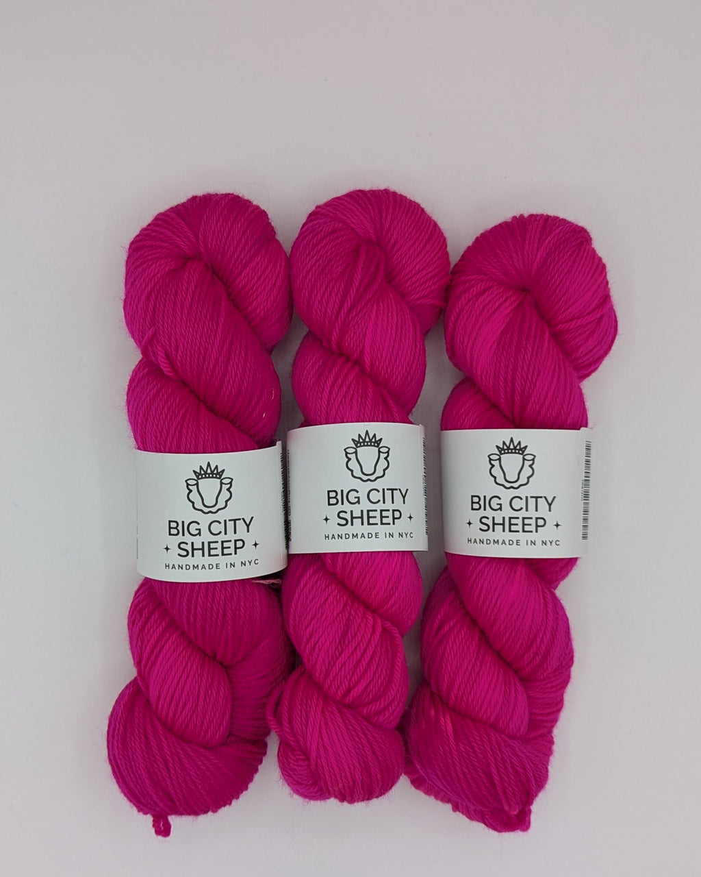 Three skeins of bright magenta pink DK weight yarn with white paper labels reading 'BIG CITY SHEEP' arranged vertically on a white surface.