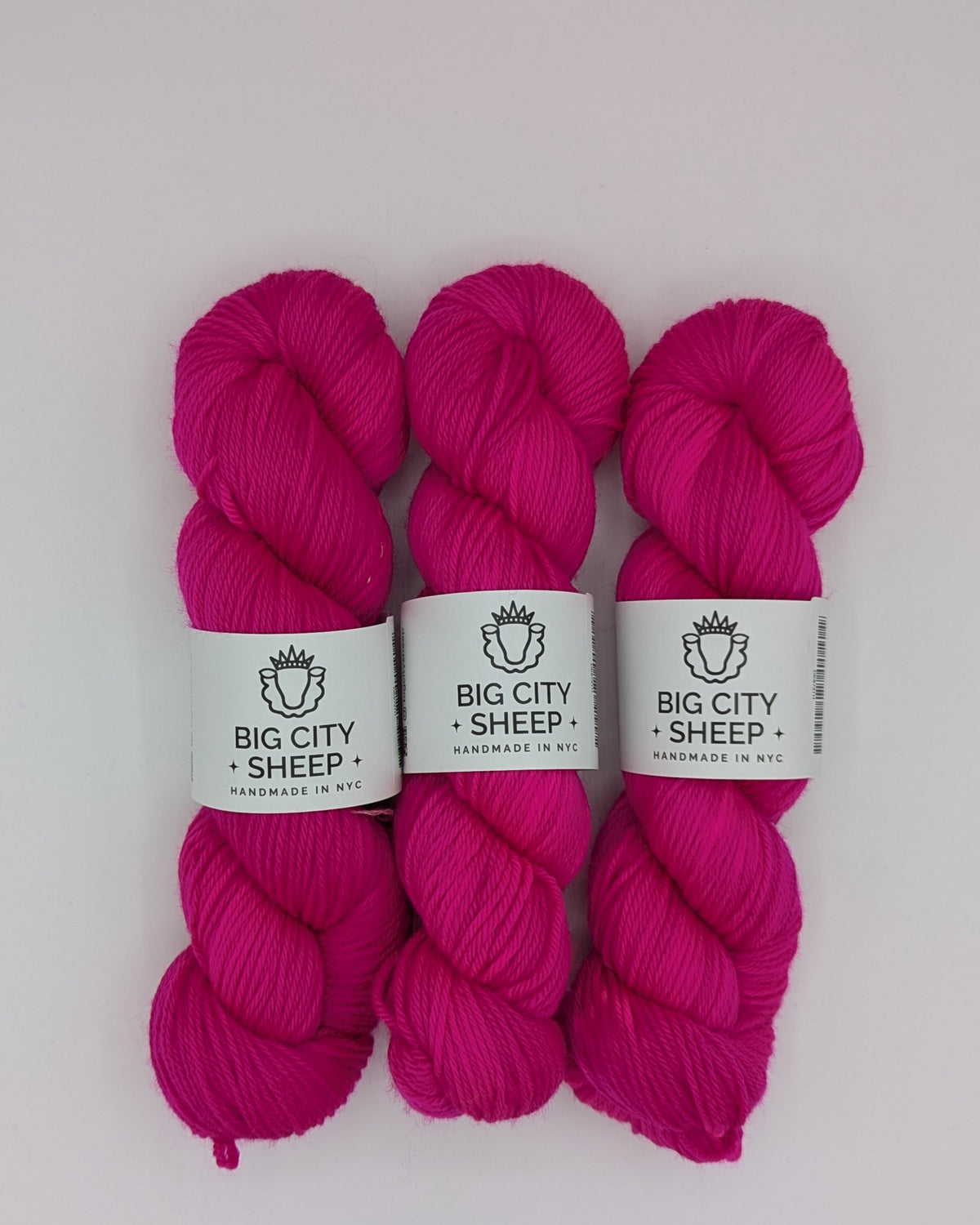 Three skeins of bright magenta pink DK weight yarn with white paper labels reading 'BIG CITY SHEEP' arranged vertically on a white surface.