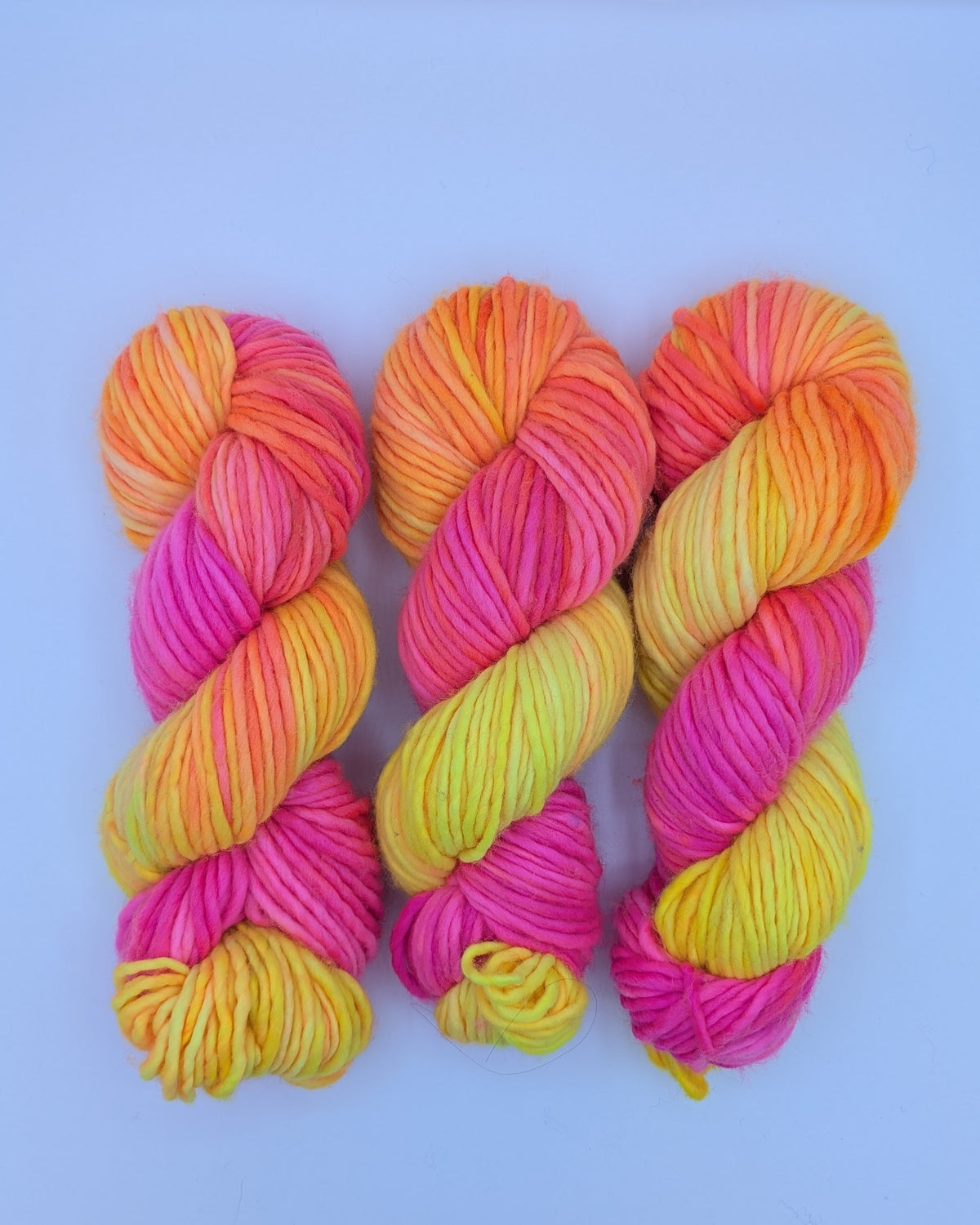 Three skeins of chunky variegated yarn in bright pink, orange, yellow, and pale green, arranged side by side on a light blue background.